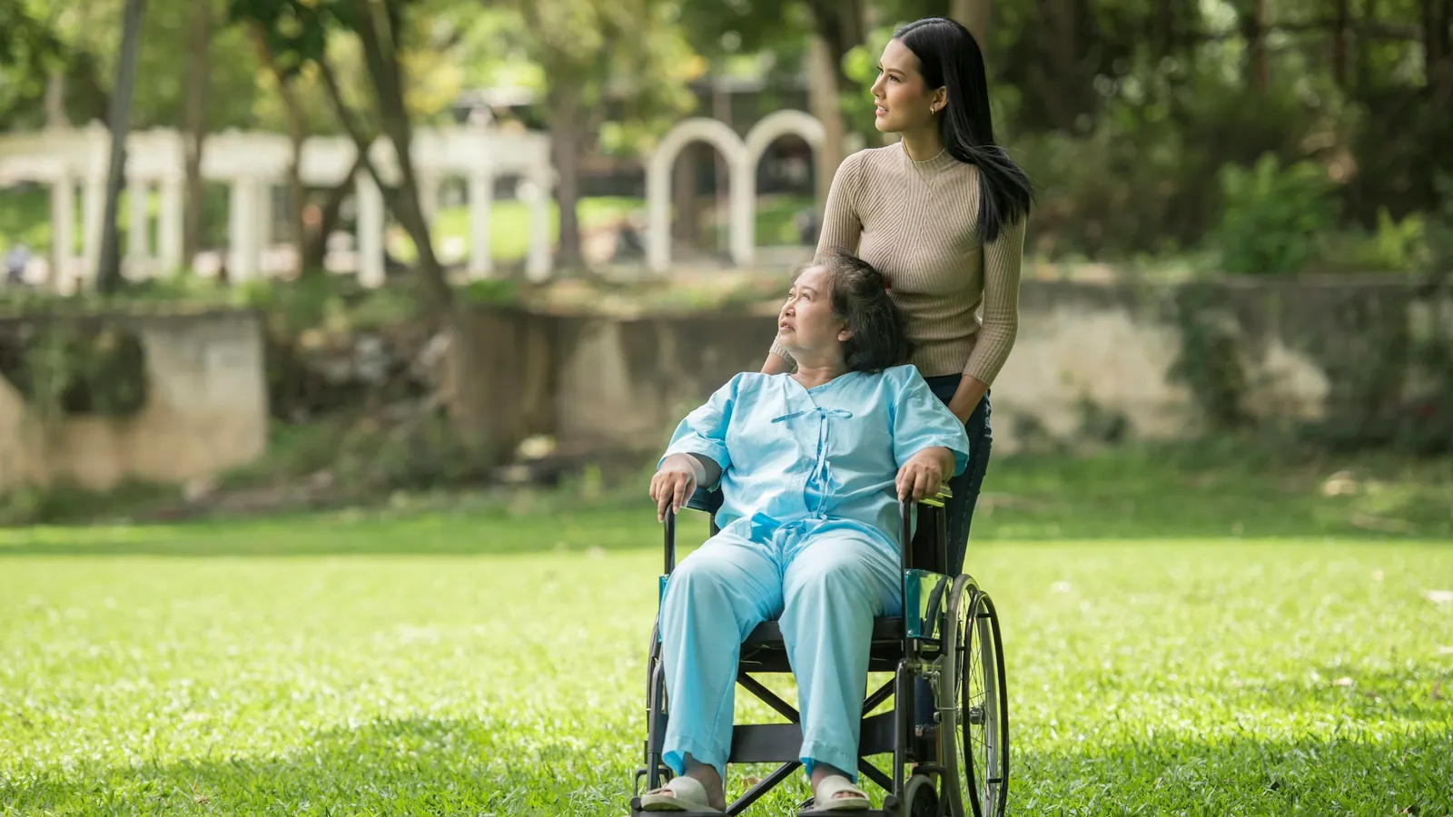 A young woman pushes an elderly woman in a wheelchair across a park lawn