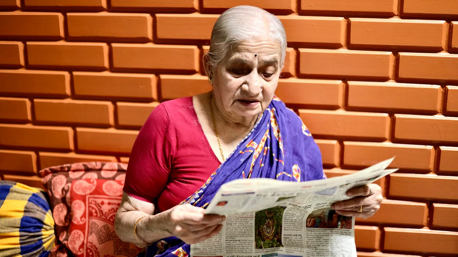 An elderly woman in a sari reads a newspaper at home