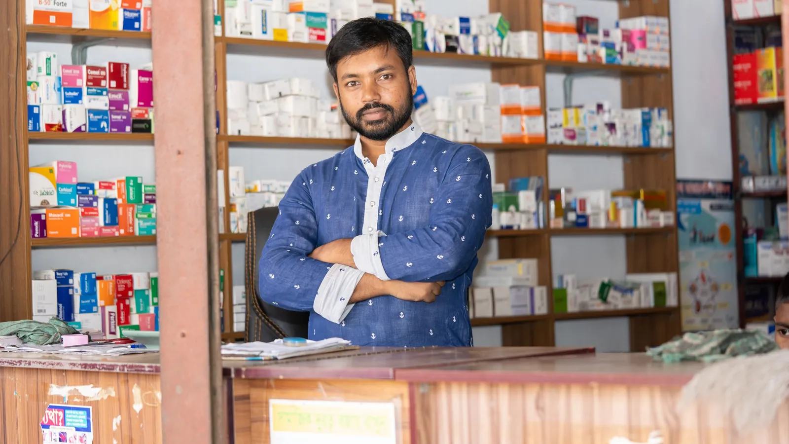 A pharmacist stands behind the counter of a pharmacy with shelves of medicines